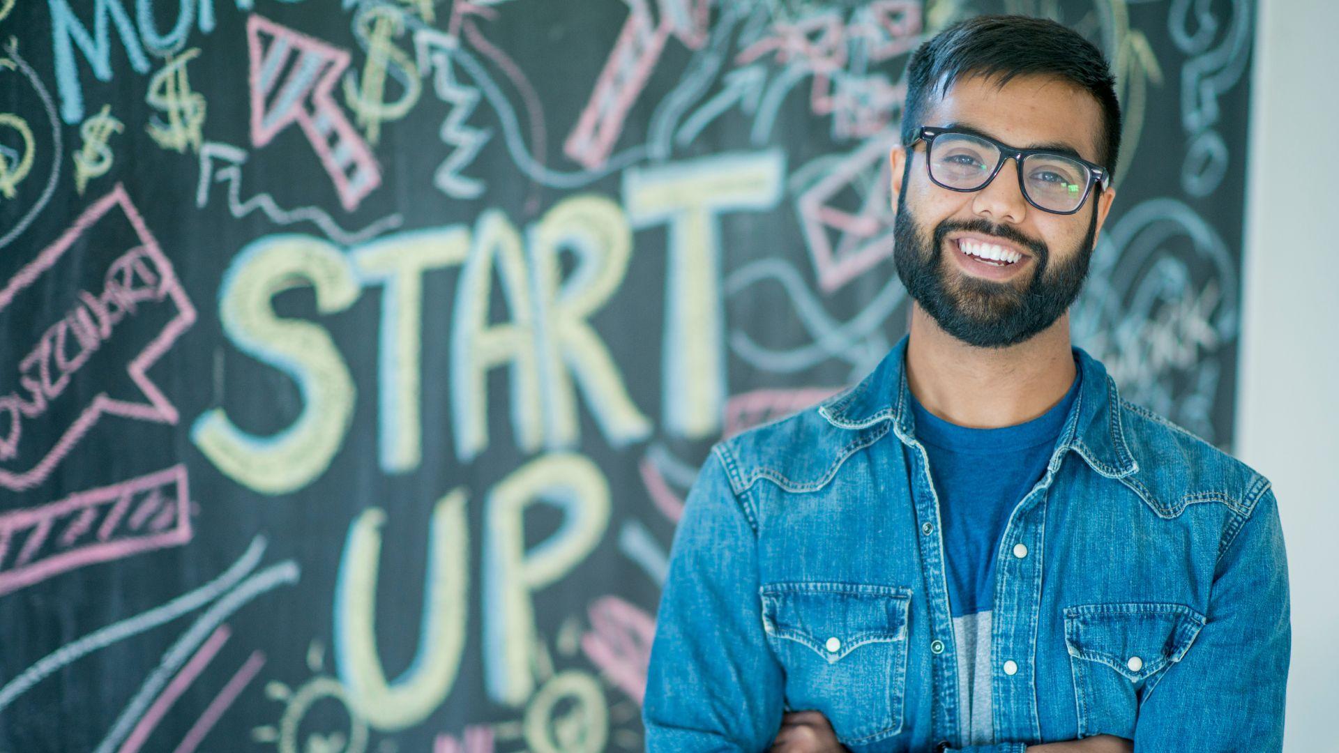 A smiling young man in front of a chalkboard that has 'Start Up' written on it